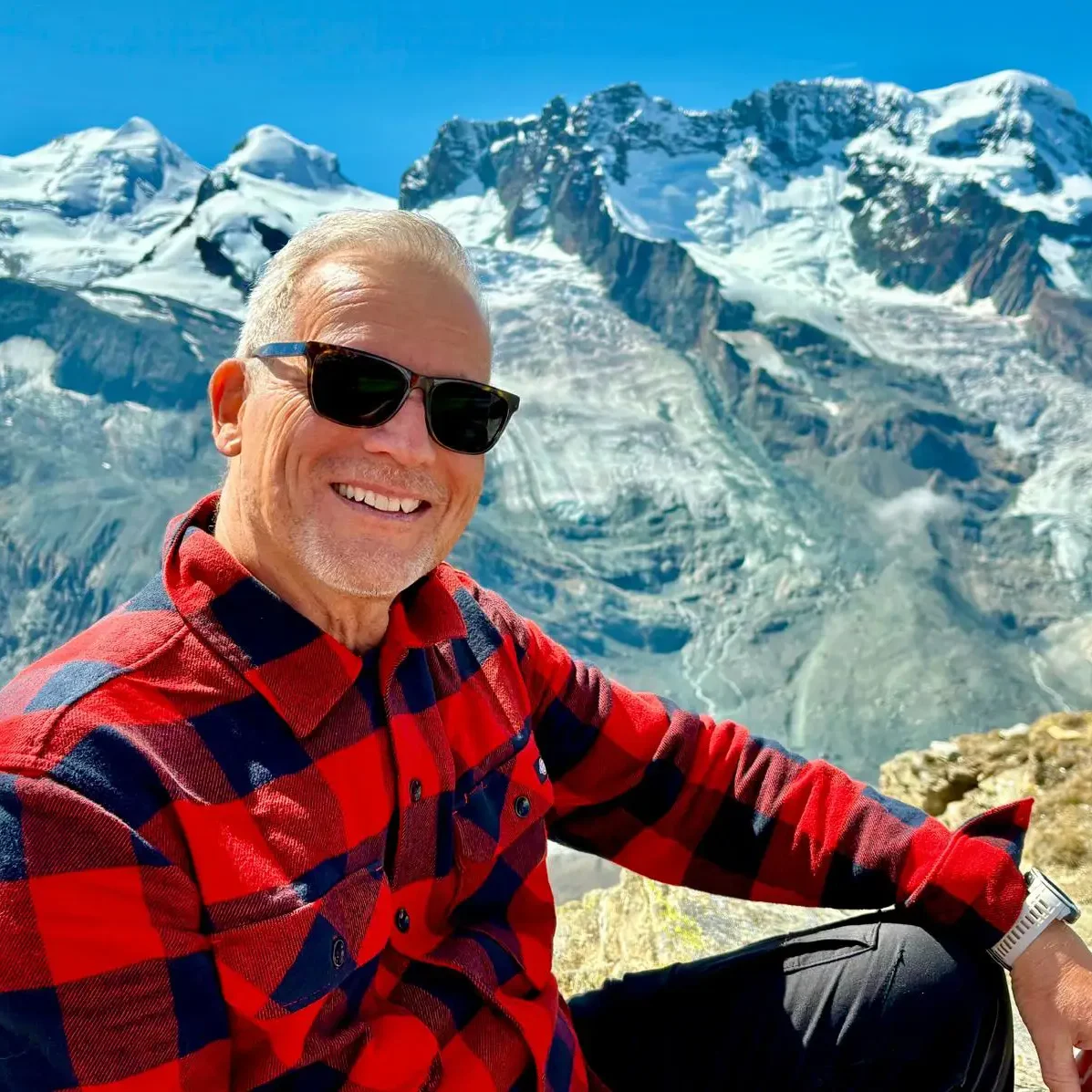 Michael Deusch Mann mit rotem kariertem Hemd und Sonnenbrille sitzt vor einer schneebedeckten Hochgebirgslandschaft mit Gletschern und blauem Himmel.