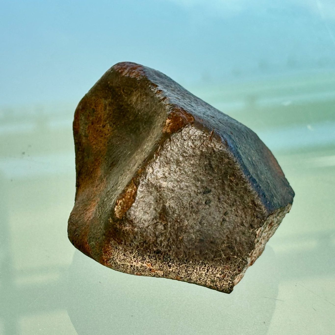 Meteorit Dunkler, kantiger Meteorit mit silbrig-metallisch glänzender Oberfläche und rostbraunen Verfärbungen vor hellem Hintergrund.